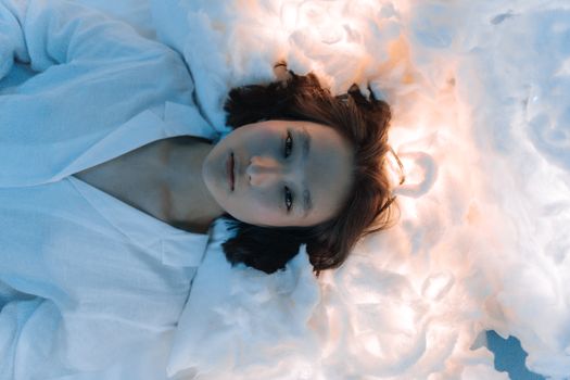 Serene portrait of an Asian woman lying on fluffy white fabric under soft lighting.