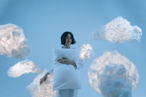 Free A serene portrait of a woman holding a pillow surrounded by soft clouds, evoking a dreamlike atmosphere. Stock Photo
