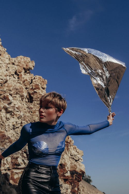 Young stylish woman with balloon in outstretched arm looking away against rough rock under cloudy blue sky