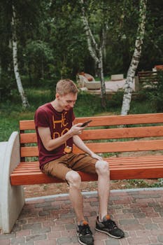 Smiling man in a red shirt using his phone while sitting on a bench outdoors.