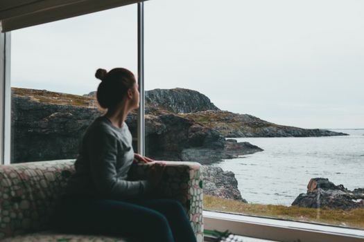 A woman sits indoors looking through a window at a rocky coastal landscape.
