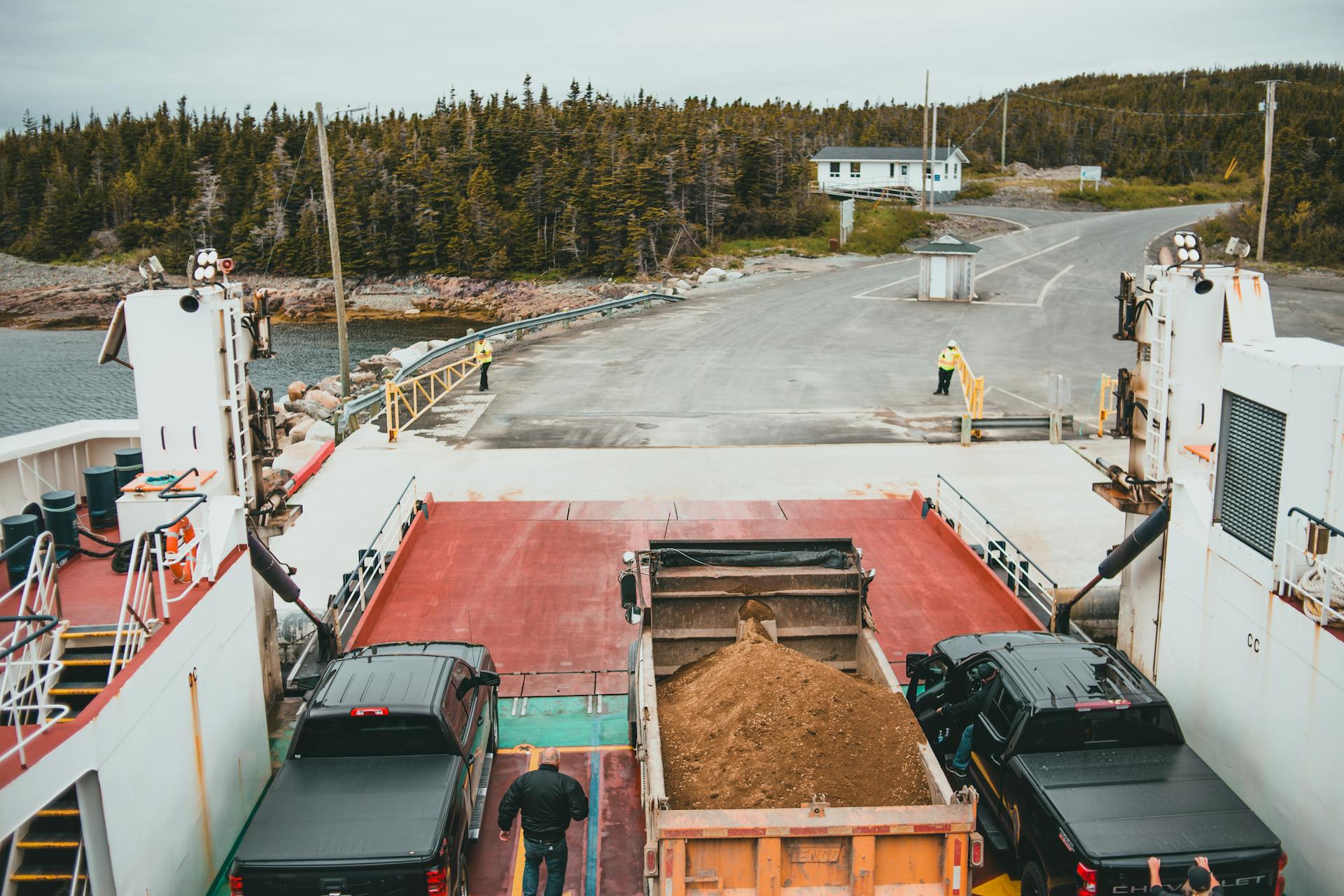 Trucks disembarking a ferry at a forested coastal location on a cloudy day.