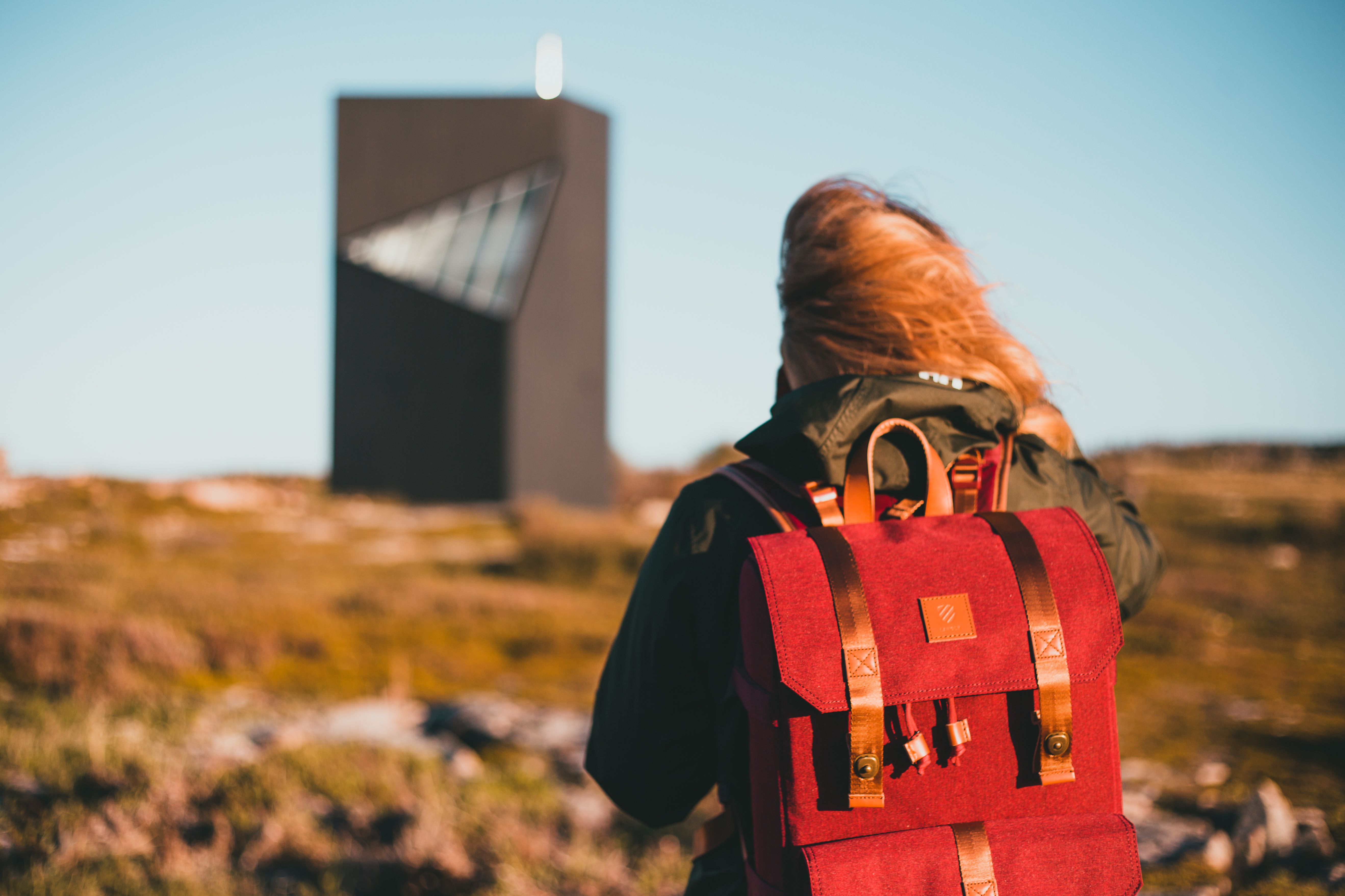 A woman with a red backpack hikes near a modern building under clear skies.