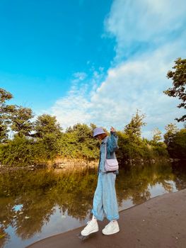 A woman in denim and bucket hat posing by a serene river with a blue sky backdrop.