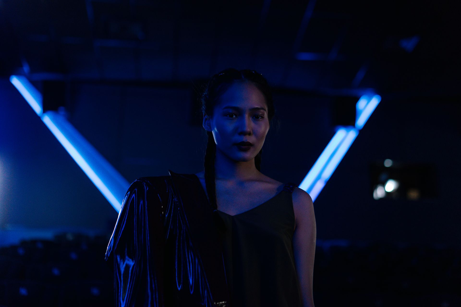 An Asian woman stands stylishly illuminated by neon blue lights indoors, creating a cinematic mood.
