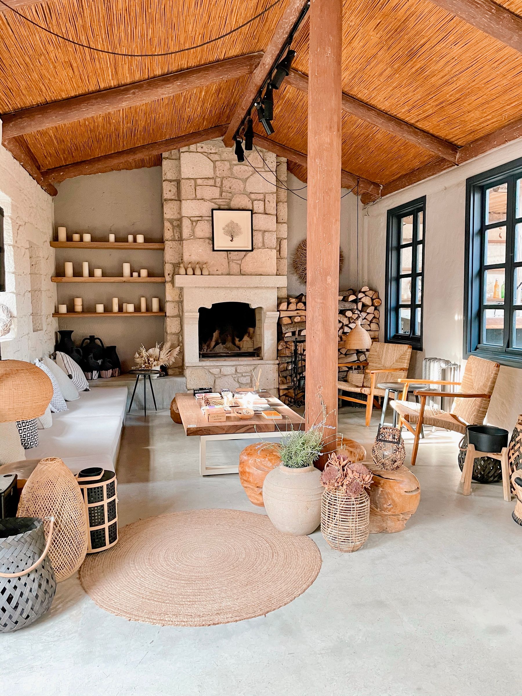 Rustic living room with a stone fireplace, timber-and-reed ceiling, woven chairs, pale sofa and stacked firewood