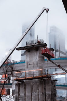 A construction site with cranes assembling a concrete bridge in an urban setting with skyscrapers in the background.