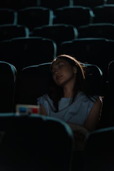 A woman peacefully sleeps in a dark movie theater holding 3D glasses, surrounded by empty seats.