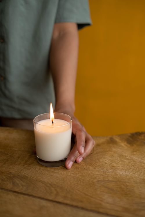 Free A warm candle held by a hand on a wooden table against a yellow background, creating a cozy ambiance. Stock Photo