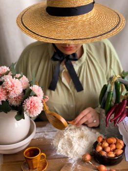 Unrecognizable female in hat using spoon for mixing flour on table near onions and hot peppers and cup near flowers