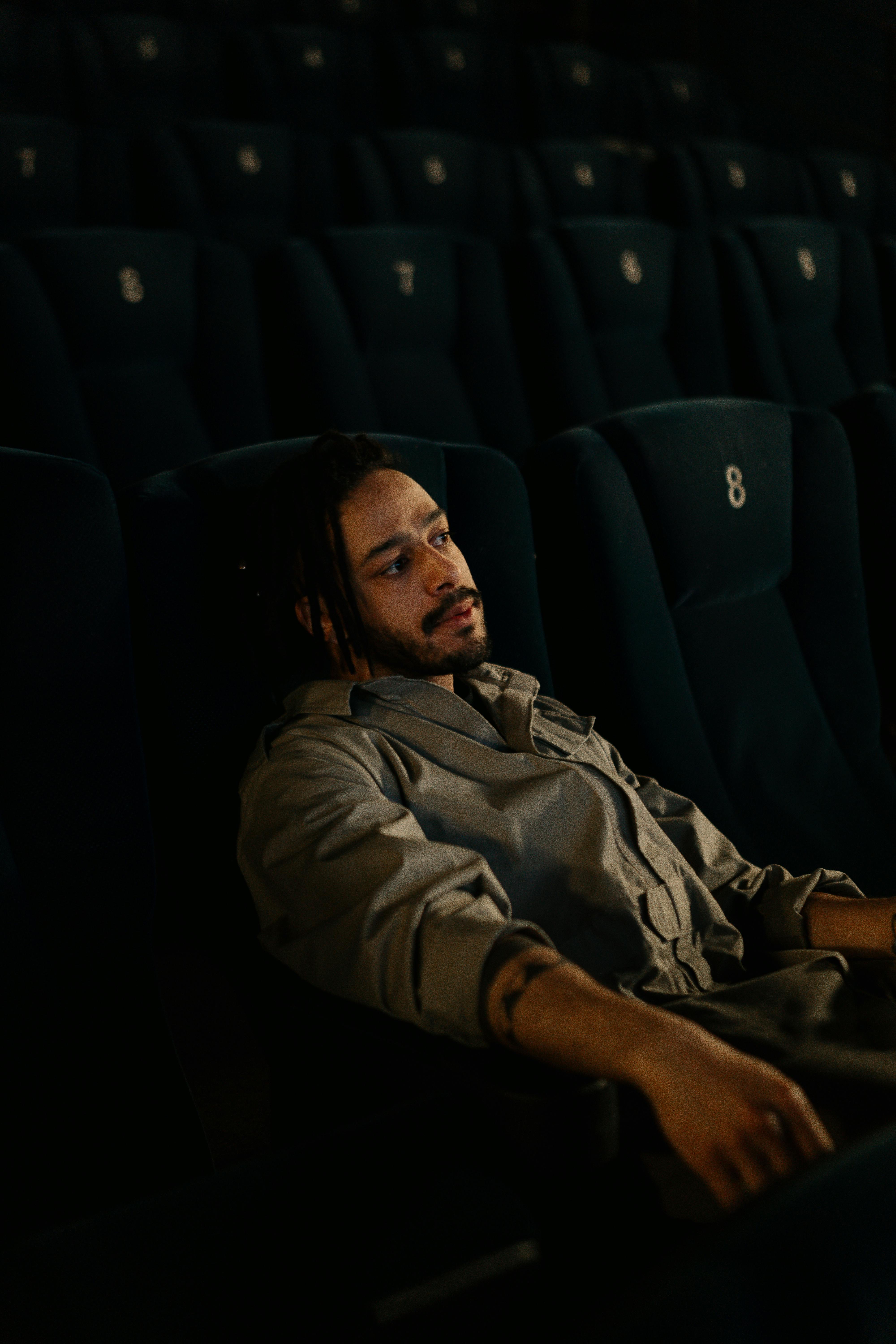 Free A man with braided hair sits thoughtfully in an empty cinema with dim lighting. Stock Photo