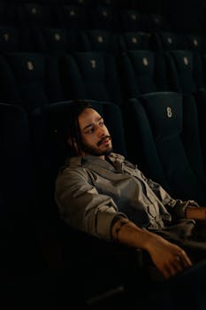 A man with braided hair sits thoughtfully in an empty cinema with dim lighting.