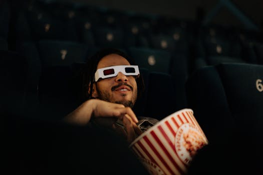 A man wearing 3D glasses enjoys popcorn while watching a movie in a cinema.