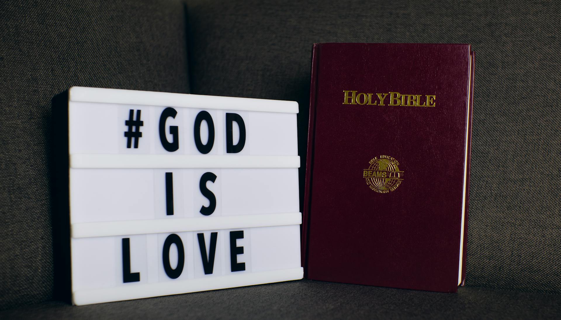 Holy Bible next to a lightbox displaying '#God is Love' message on a sofa.