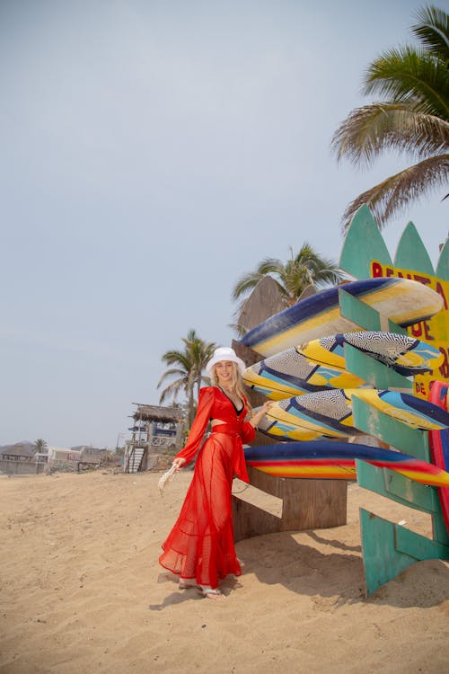 A Woman in Red Dress Standing Beside a Rack of Surfboards on the Beach