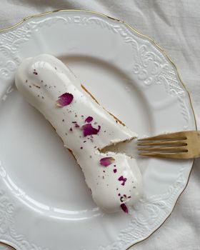 A delicious eclair topped with cream and flower petals on a ceramic plate with a golden fork.