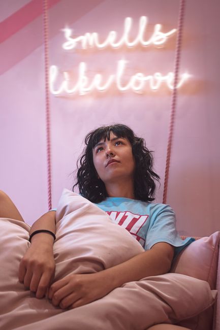 Woman with curly hair relaxes indoors, gazing at a neon sign reading 'smells like love'.