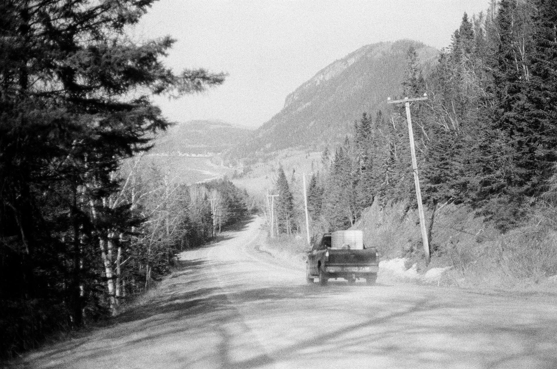 Black and white image of a truck driving on a rural mountain road lined with trees.