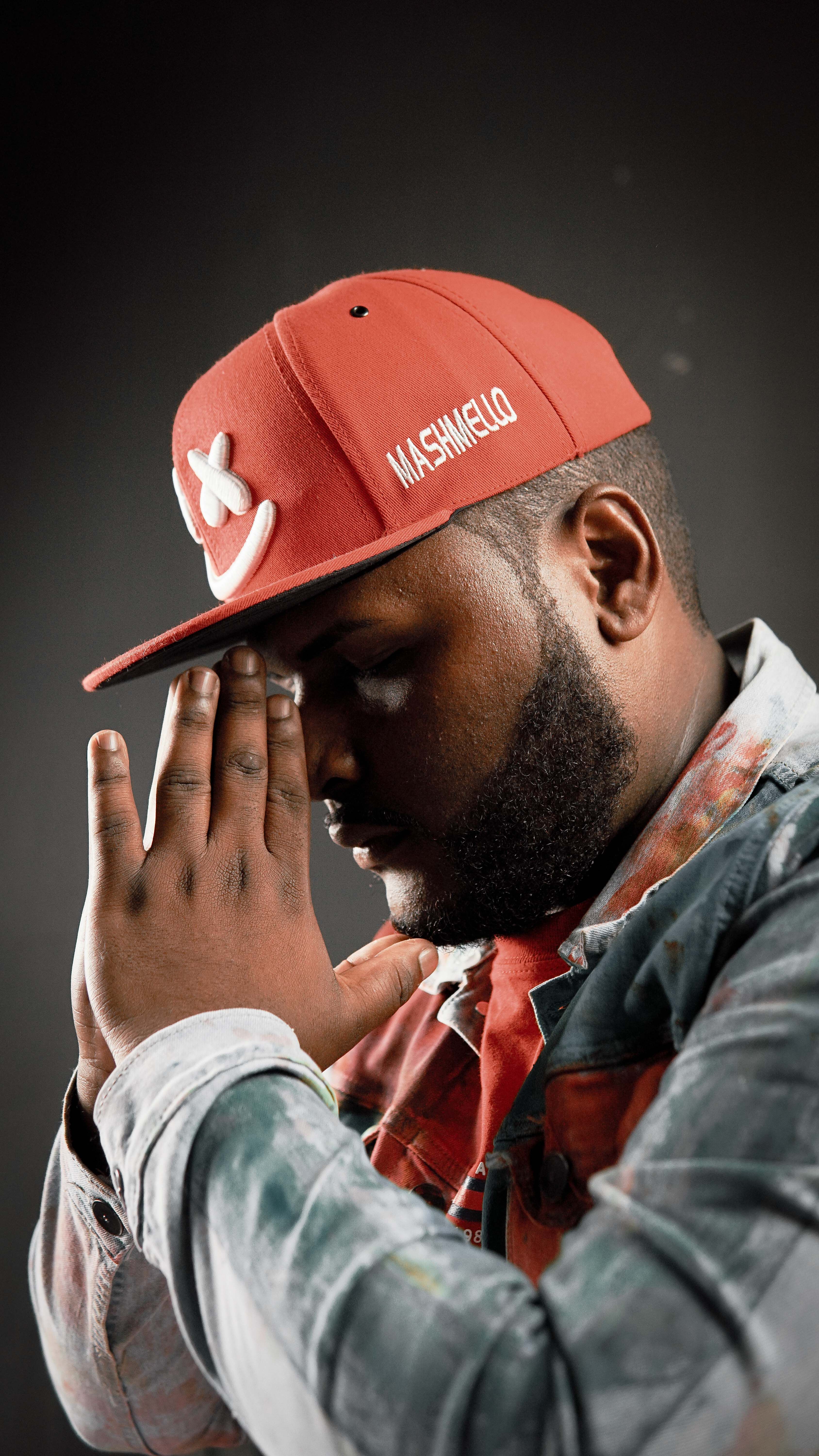 Side profile of a man in a red cap, hands clasped in thought against a dark background.