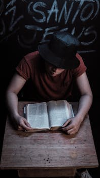 Young man wearing hat reading a Bible at a wooden desk in a dimly lit room with religious graffiti.