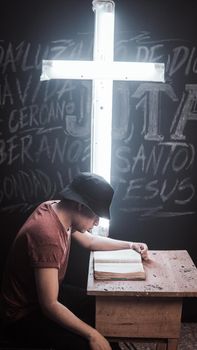 A young man wearing a black hat reads a book by an illuminated cross in a dimly lit room.