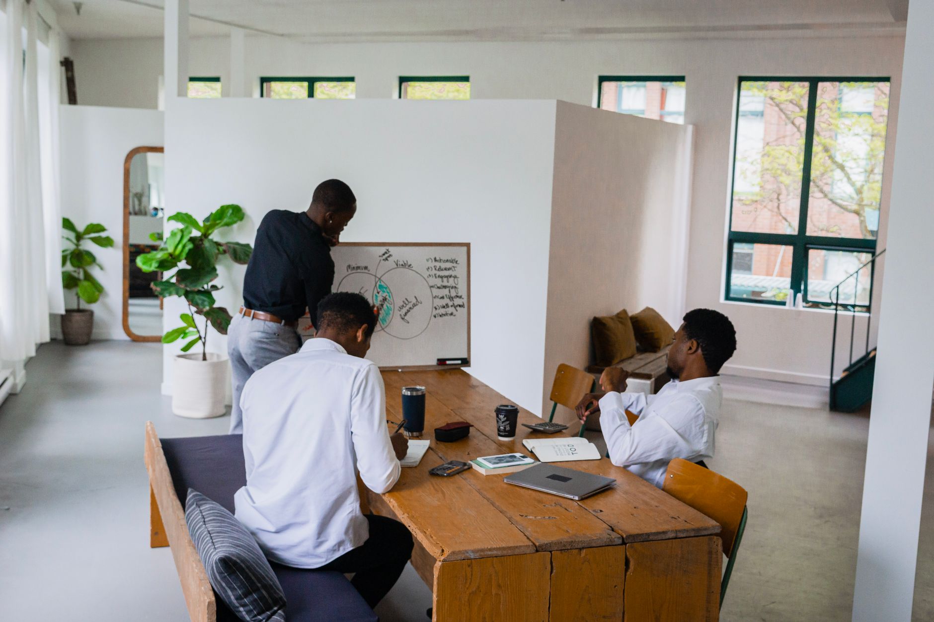 Three men engage in a strategy meeting with a whiteboard in a bright, green-filled office space.