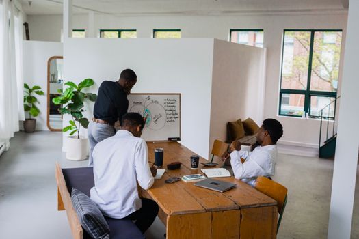 Three men engage in a strategy meeting with a whiteboard in a bright, green-filled office space.