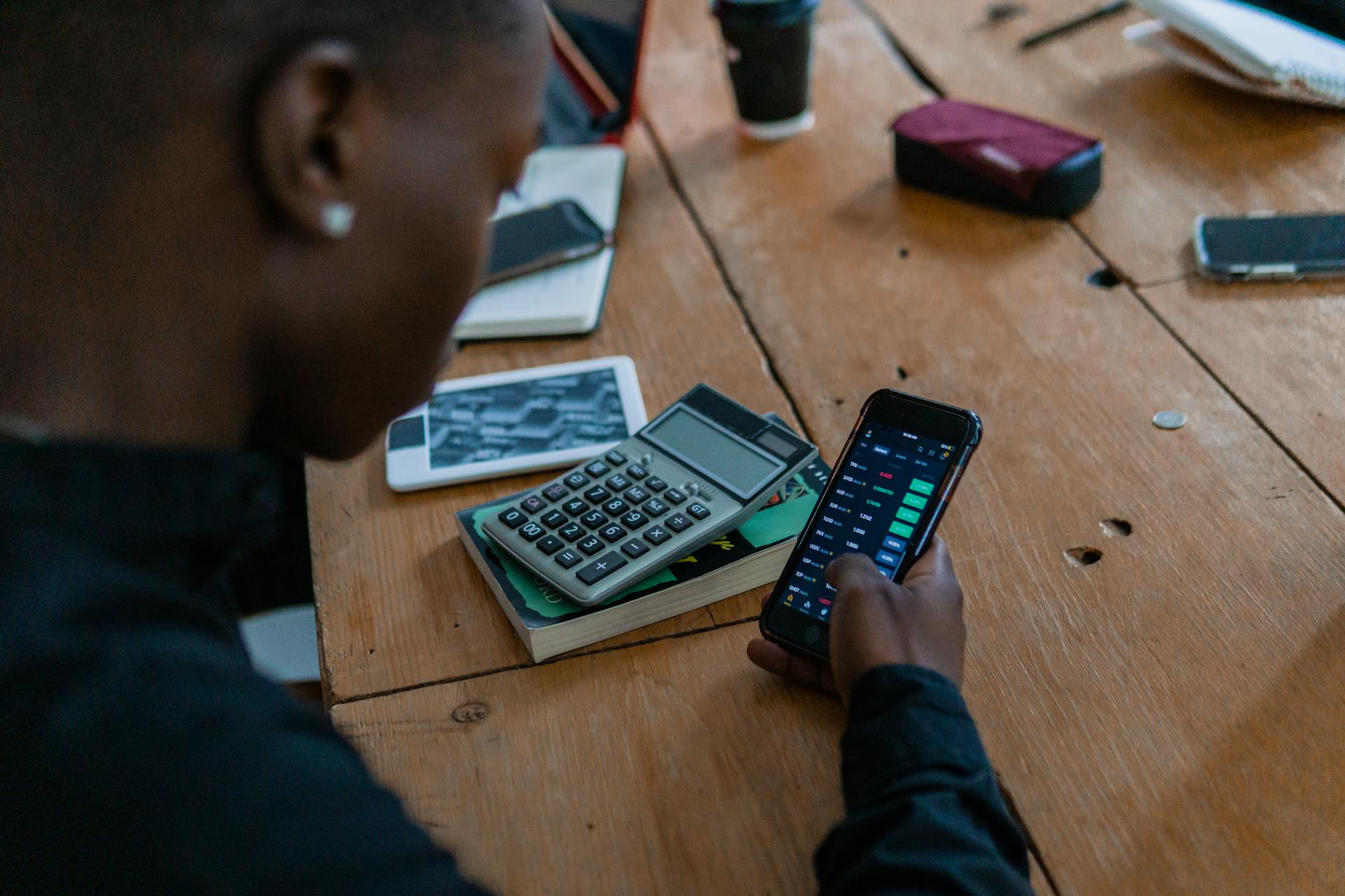 Close-up of individual using smartphone for financial data analysis, calculator nearby on wooden table.