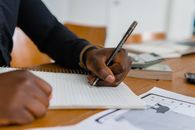 Close-up of hand writing on notebook at a wooden table, featuring paper and ballpen.