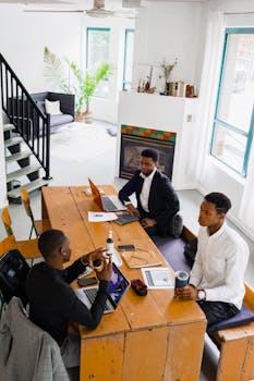 Three colleagues engaged in a discussion around a wooden table in a bright office setting.