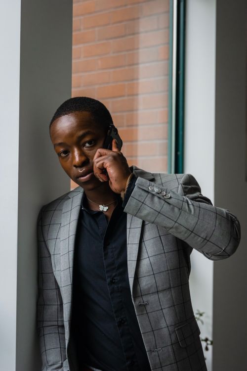Free African American man in a gray checkered suit making a phone call indoors, leaning against a window. Stock Photo