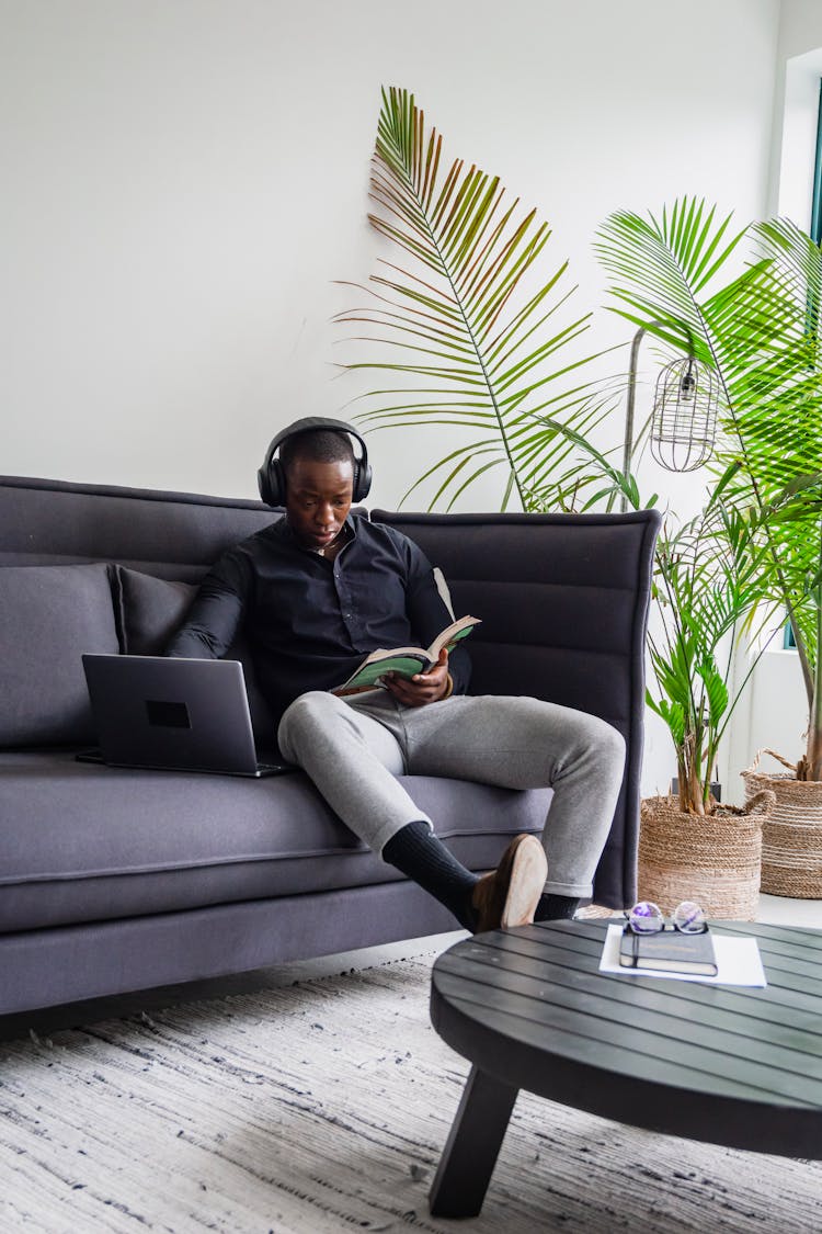 A Man Reading A Book Beside His Laptop