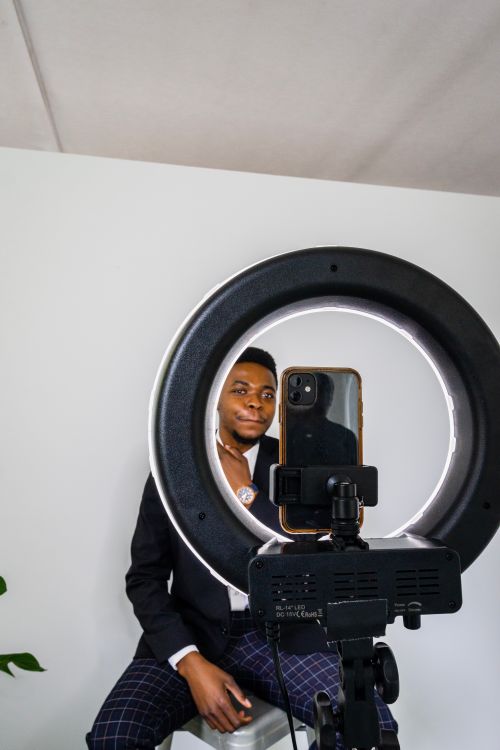 Free Man in black suit using a smartphone ring light for professional headshot, seated indoors. Stock Photo