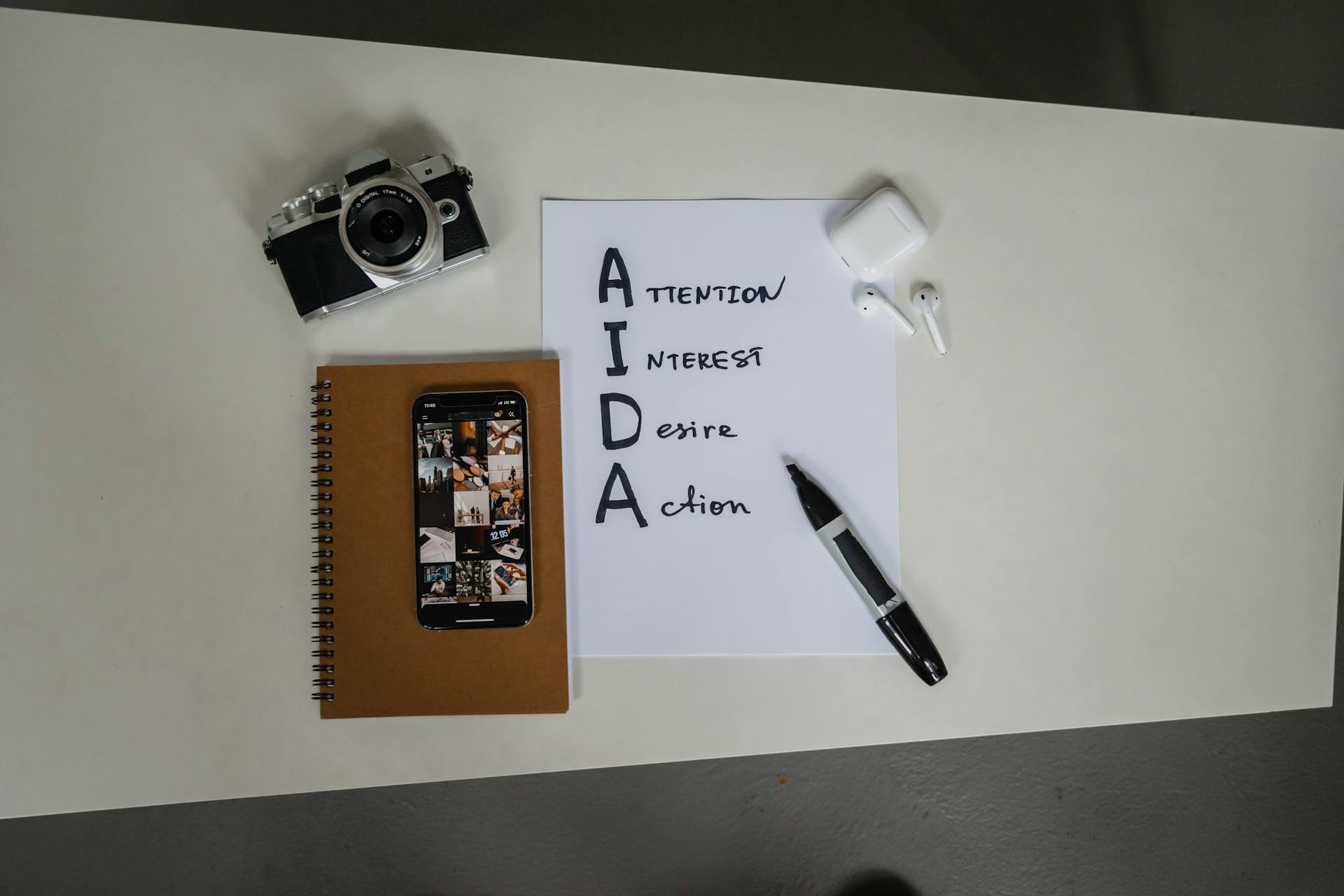 Top view of AIDA marketing elements with notebook, smartphone, camera, and earbuds on a desk.