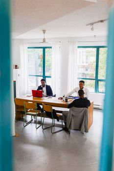Three businessmen engaged in a meeting in a bright, modern office space with large windows.