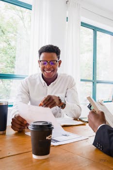 Smiling man reviewing documents in a brightly lit office with coffee and colleagues.