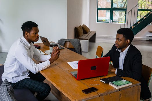 Two men in a business meeting at a wooden table, discussing with laptops and notes.