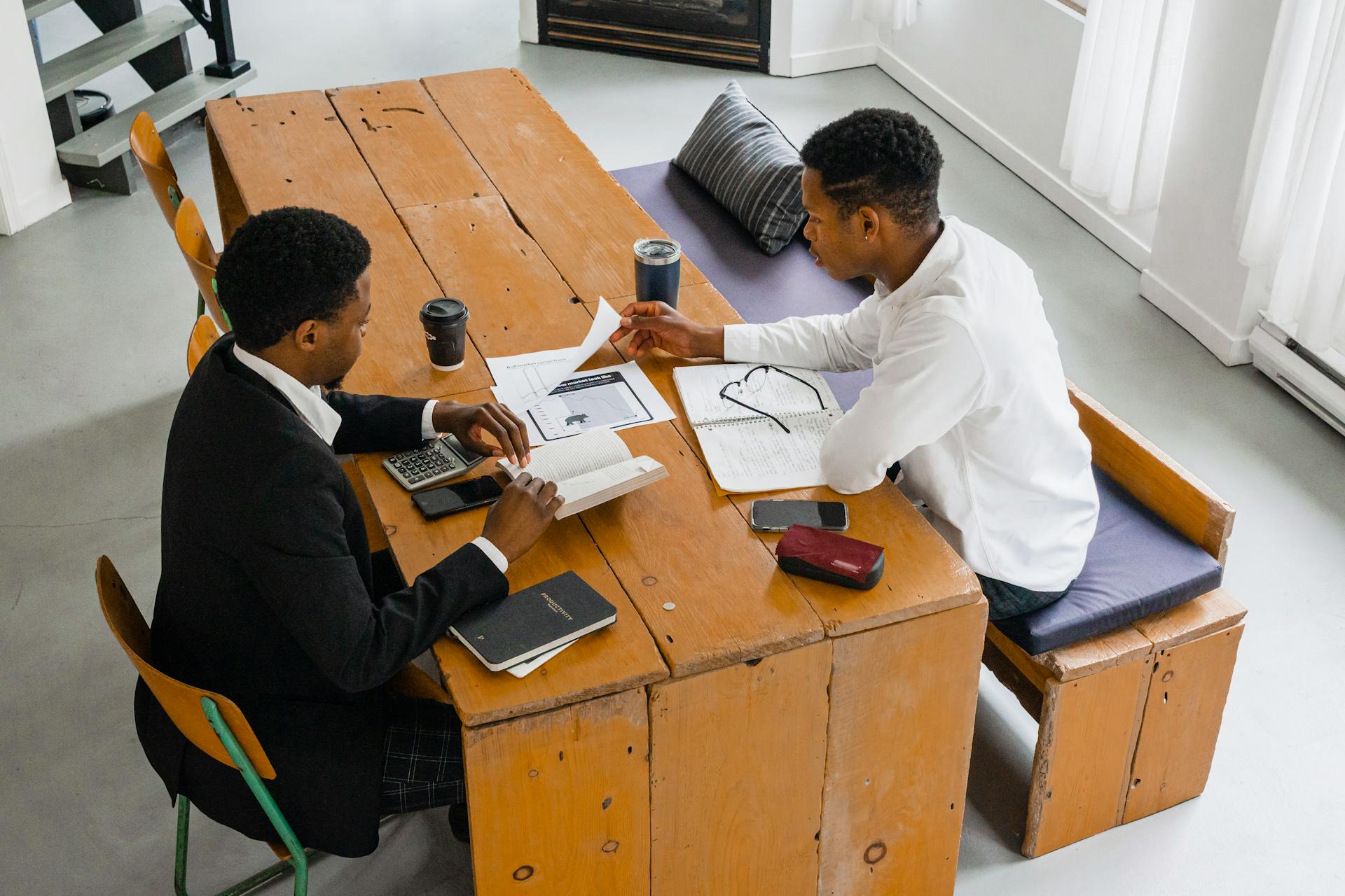 Two men discuss finances in a modern office setting, reviewing documents and notebooks.