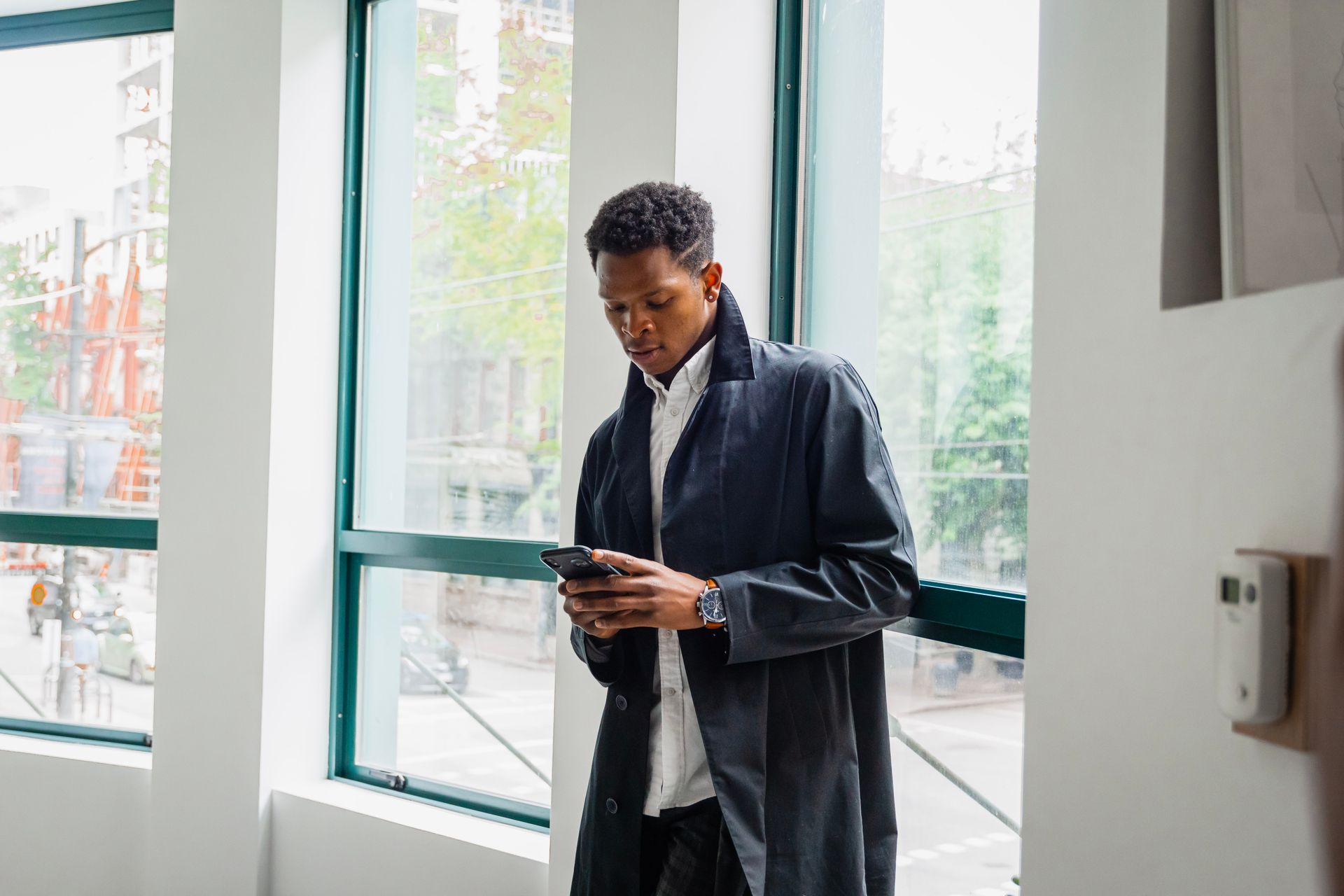 Businessman in a black jacket using smartphone indoors by office window. Modern and professional setting.