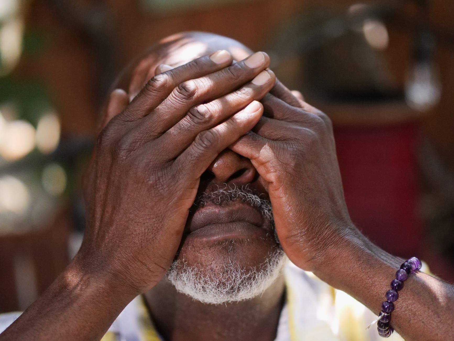 Close-up of an elderly man with a beard covering his face with his hands outdoors.