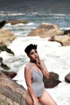 Adult woman in a striped swimsuit posing by the rocky seashore, suggesting a summer day at the beach.