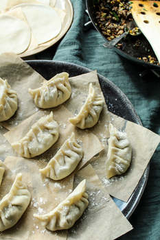 Raw dumplings on parchment paper with dough discs and filling ingredients nearby.