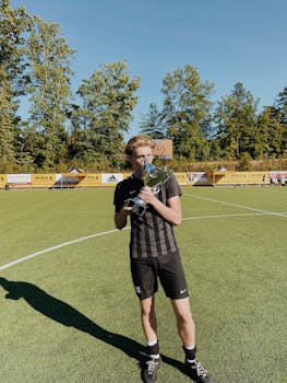 A young male soccer player proudly holding a trophy on a sunny day at the soccer field.