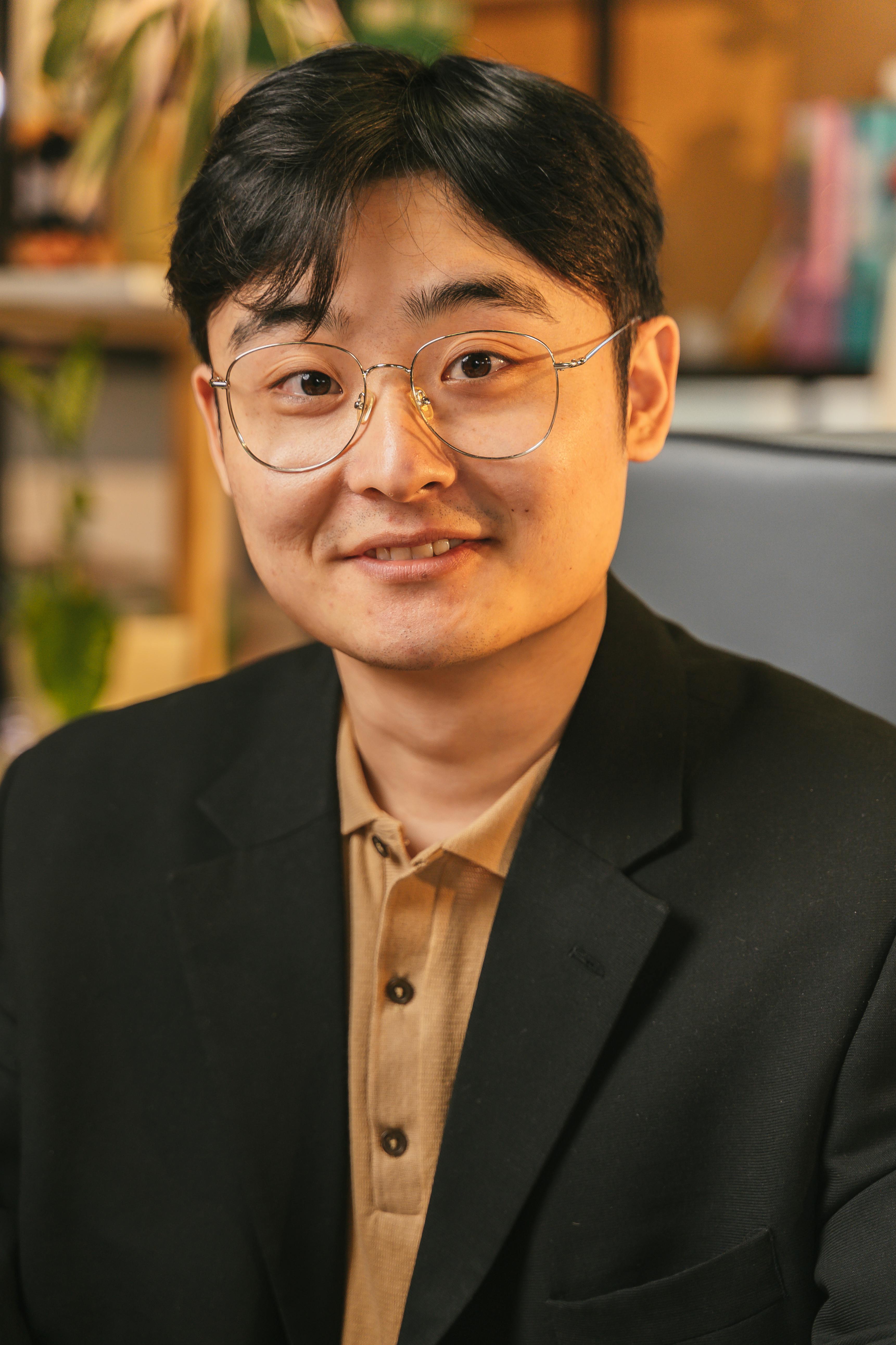 Casual portrait of a smiling man with glasses indoors, warm lighting accentuates friendly expression.