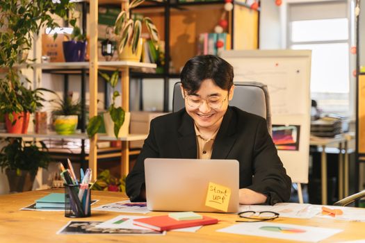 Smiling young professional in black suit working on laptop at a cozy office desk surrounded by plants.