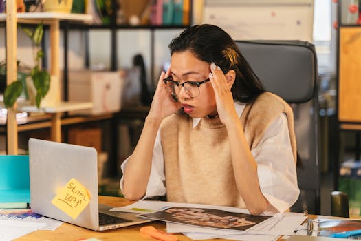 A woman with glasses shows stress while working at a laptop in an office environment.