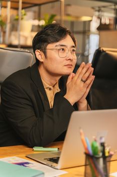 Adult man in black suit thoughtfully working on laptop in modern office setting.