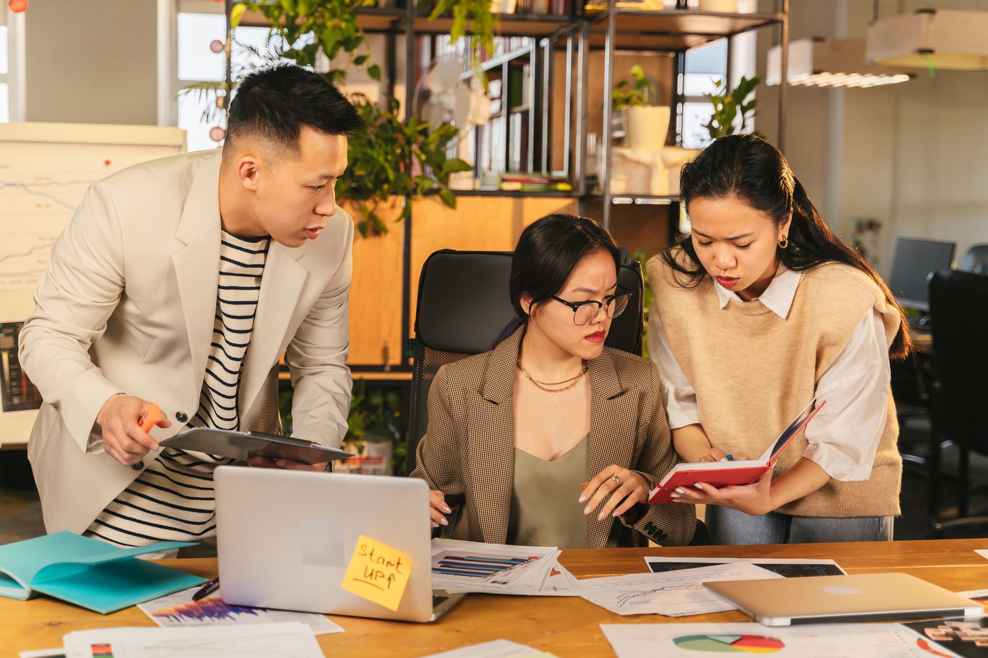 Team of professionals engaged in a discussion around a table with laptops and documents in a modern office environment.