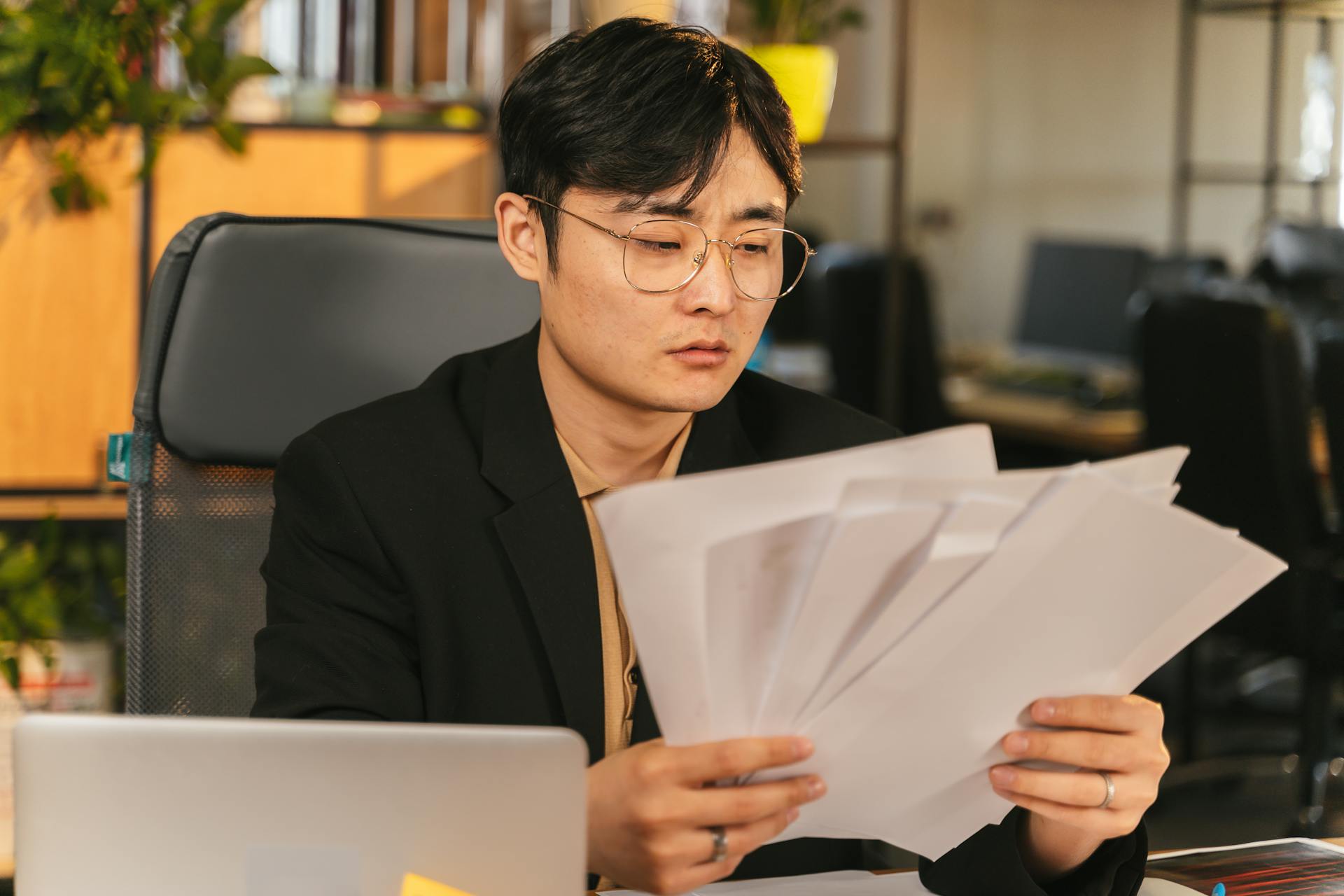 Asian man in glasses and suit analyzing papers at his desk in a modern office.