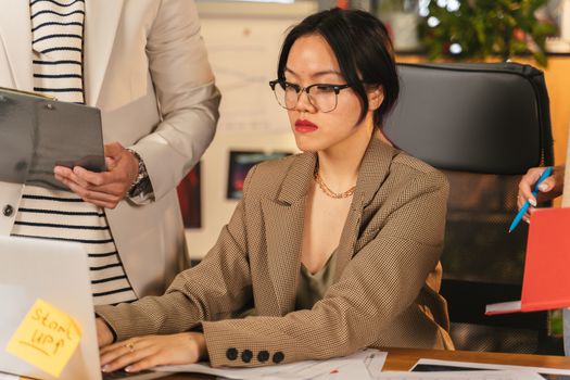 Professional Asian woman in glasses working on a laptop in an office environment.